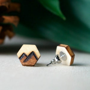 May include: Hexagon-shaped wooden stud earrings. Each earring features a mountain range design in dark brown against a light wood background. The earrings have silver posts and are set against a soft, neutral background.