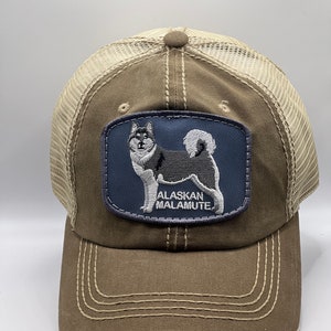 Puede incluir: Una gorra de camionero marrón y beige con un aspecto desgastado. La gorra tiene un parche azul con una imagen bordada de un perro Alaskan Malamute y el texto "Alaskan Malamute".