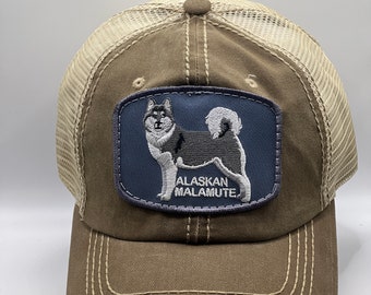 May include: A brown and beige trucker hat with a distressed look. The hat features a blue patch with an embroidered image of an Alaskan Malamute dog and the text "Alaskan Malamute".