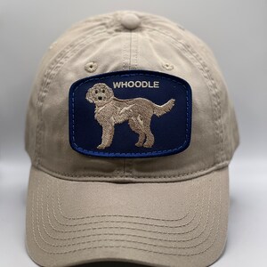 May include: Khaki baseball cap with a navy blue patch featuring an embroidered image of a dog and the word "WHOODLE" in white. The cap has a curved brim and a button on top.