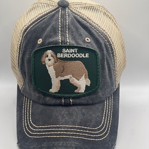 May include: A black and beige trucker hat with a distressed look. The hat features a green patch with a brown and white Saint Berdoodle dog and the text "SAINT BERDOODLE".