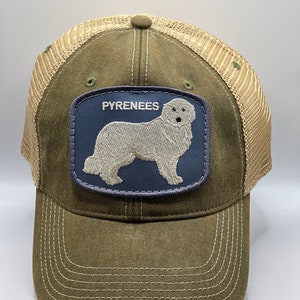 Puede incluir: Gorra de béisbol verde oliva con parte trasera de malla y visera curva. Parche azul marino con la palabra "PYRENEES" y un bordado de un perro de los Pirineos.