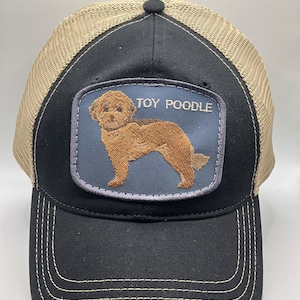 May include: A black and tan trucker hat with a mesh back. The hat has a patch on the front that features a brown toy poodle with the text "TOY POODLE" above it.