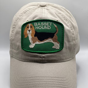 Könnte beinhalten: Eine beige Baseballkappe mit einem gestickten grünen Aufnäher, der einen Basset Hound und den Text "BASSET HOUND" zeigt.