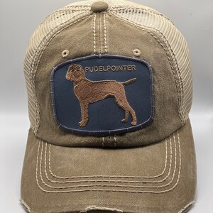 Op de afbeelding: Een bruine en beige verweerde truckercap met een blauwe patch met een geborduurd bruin hondensilhouet en de tekst "PUDELPOINTER".