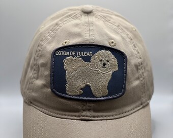 May include: A beige baseball cap with a navy blue patch featuring a white Coton de Tulear dog and the text "Coton de Tulear".