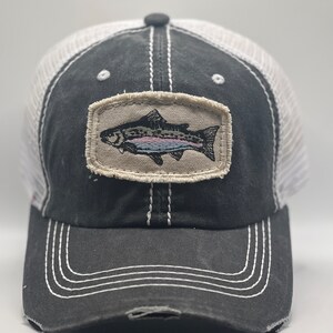 May include: A black and white trucker hat with a distressed look. The hat features a brown patch with an embroidered image of a trout fish.