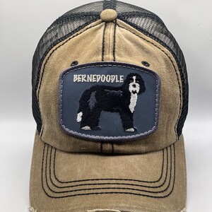 May include: A brown and black trucker hat with a black mesh back. The hat has a distressed look and features a patch with a black and white Bernedoodle dog and the text "BERNEDoodle".