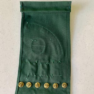 May include: Dark green fabric with embroidered details and gold-colored buttons. The fabric has a stitched circular design and curved lines, with several small stitches below. Six gold buttons are visible at the bottom of the fabric.