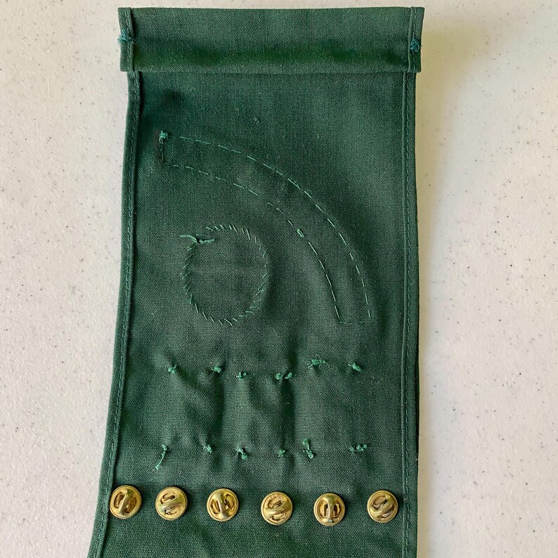 May include: Dark green fabric with embroidered details and gold-colored buttons. The fabric has a stitched circular design and curved lines, with several small stitches below. Six gold buttons are visible at the bottom of the fabric.