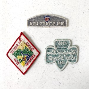 May include: Three embroidered patches: a diamond-shaped patch with a landscape scene, a trefoil patch, and a rectangular patch with the text "GIRL SCOUTS USA". The patches are in various colors including red, green, yellow, and white.