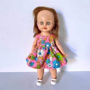 May include: A vintage doll with auburn hair and large blue eyes. The doll wears a pink dress with a floral pattern in white, blue, and green. White shoes complete the outfit. The doll is standing against a plain white background.