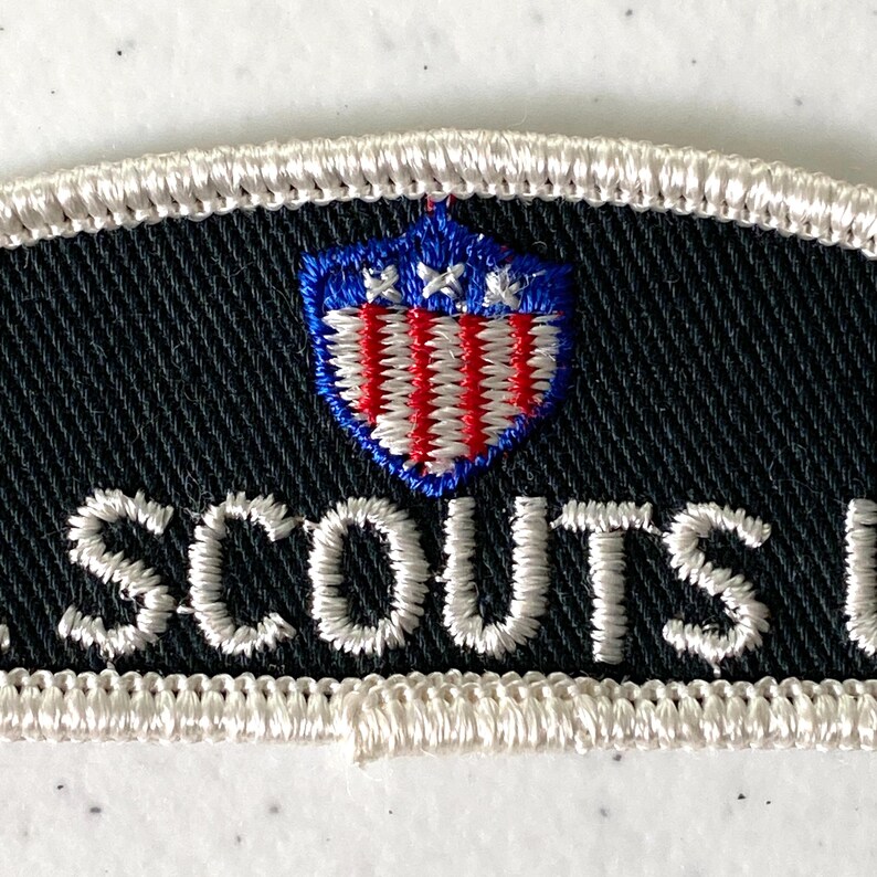 May include: Embroidered patch with the word "SCOUTS" in white thread on a black background. The patch features a shield design in red, white, and blue, with a white border.