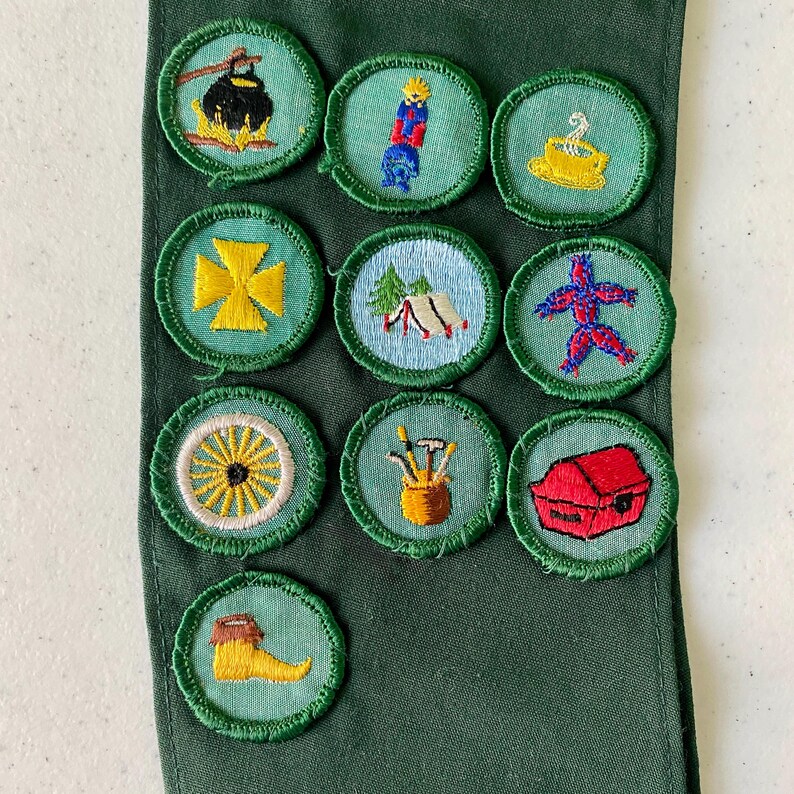 May include: A collection of embroidered patches with various designs, including a campfire, a figure, a tent, and a wagon wheel. The patches are circular with green borders and are attached to a green fabric.