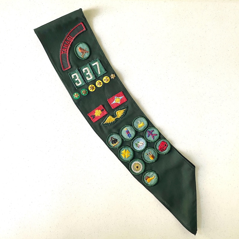 May include: Dark green sash with various embroidered patches. Patches include the number "337", a bird, stars, and other colorful designs. The word "TANASI" is embroidered in red on a curved patch.