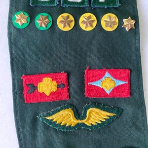 May include: A collection of military patches and pins on a dark green fabric. The patches are red with yellow and blue designs. Gold star-shaped pins and green and yellow circular pins are also visible.