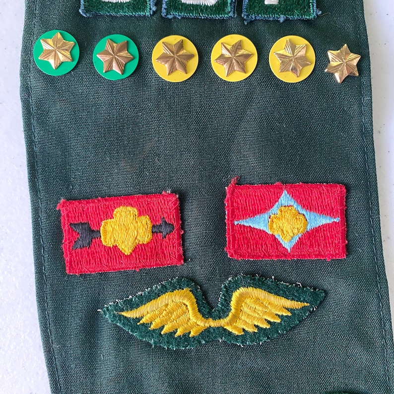 May include: A collection of military patches and pins on a dark green fabric. The patches are red with yellow and blue designs. Gold star-shaped pins and green and yellow circular pins are also visible.
