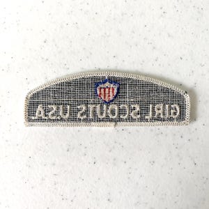 May include: A rectangular Girl Scouts USA patch with a white border and a black mesh background. The patch features a shield emblem in red, white, and blue, and the words "GIRL SCOUTS USA" in white.