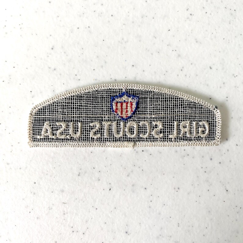 May include: A rectangular Girl Scouts USA patch with a white border and a black mesh background. The patch features a shield emblem in red, white, and blue, and the words "GIRL SCOUTS USA" in white.