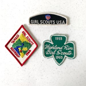 May include: Three vintage Girl Scout patches. One black patch with "Girl Scouts U.S.A." text. A red and white diamond-shaped patch with "Tanasi Girl Scouts Roundup 1966". A green shamrock-shaped patch with "Highland Rim Girl Scouts 1955 1965".
