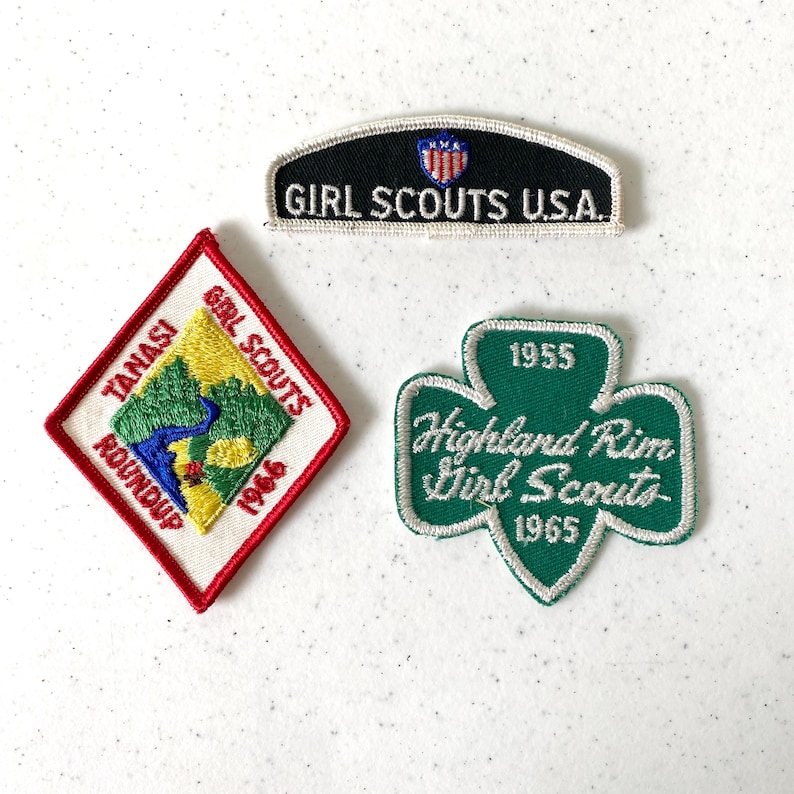 May include: Three vintage Girl Scout patches. One black patch with "Girl Scouts U.S.A." text. A red and white diamond-shaped patch with "Tanasi Girl Scouts Roundup 1966". A green shamrock-shaped patch with "Highland Rim Girl Scouts 1955 1965".