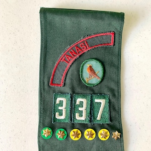 May include: Dark green fabric with embroidered patches. Includes a red arched patch with "TANASI" in white, a circular patch with a bird, and the numbers "337". Several gold and green star-shaped pins are also visible.