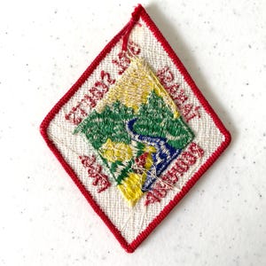 May include: Embroidered diamond-shaped patch with a red border. The patch features a landscape scene with green trees, a blue river, and yellow accents. Red text is embroidered around the edges.