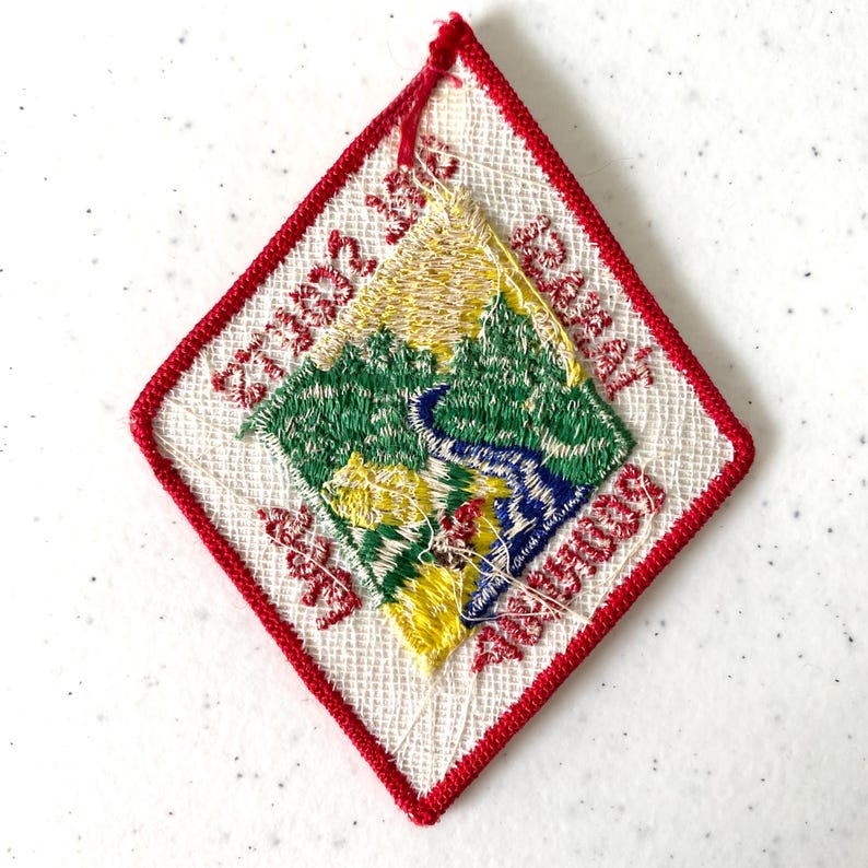May include: Embroidered diamond-shaped patch with a red border. The patch features a landscape scene with green trees, a blue river, and yellow accents. Red text is embroidered around the edges.
