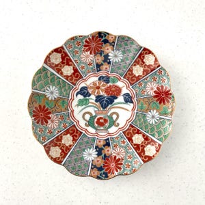 May include: Decorative ceramic plate with a scalloped edge and a floral design. The plate features a central vase with flowers, surrounded by red, green, and gold geometric patterns. The rim is trimmed with gold.