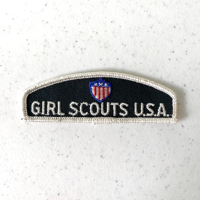 May include: Embroidered patch with the text "GIRL SCOUTS U.S.A." in white on a black background. The patch has a white border and a small emblem with red, white, and blue colors.