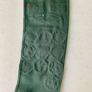 May include: A dark green fabric item with embroidered details. The top features two square shapes with red stitching and a leaf-like design. Below are several stitched circles in green thread, creating a textured pattern. The item appears to be handmade.