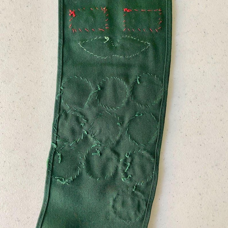 May include: A dark green fabric item with embroidered details. The top features two square shapes with red stitching and a leaf-like design. Below are several stitched circles in green thread, creating a textured pattern. The item appears to be handmade.