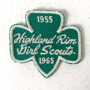 May include: A green and white embroidered Girl Scout patch in a trefoil shape. The patch features the text "Highland Rim Girl Scouts" in white script, with the years "1955" and "1965" also in white. The patch has a white border.