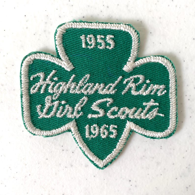 May include: A green and white embroidered Girl Scout patch in a trefoil shape. The patch features the text "Highland Rim Girl Scouts" in white script, with the years "1955" and "1965" also in white. The patch has a white border.