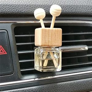 Puede incluir: Ambientador de coche con un frasco de vidrio transparente, una tapa de madera y dos difusores de bola de madera. El ambientador está colocado en una rejilla de ventilación del coche.