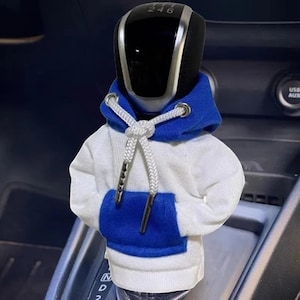 Op de afbeelding: Een zwarte en zilveren auto versnellingspookknop is gekleed in een miniatuur witte hoodie met blauwe accenten. De hoodie heeft een blauwe capuchon en zak, met een wit trekkoord. De versnellingspookknop heeft de nummers 2, 4 en 6.