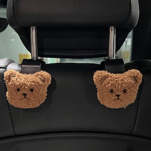 May include: Two brown teddy bear head car seat hooks. The plush bear heads have embroidered black eyes and noses. The hooks attach to the headrest of a car seat. The car seat is black.