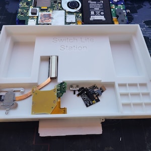 May include: White Switch Lite Station with the text "Switch Lite Station" printed on the front. The station has compartments for components, including a circuit board, and a small black module. The background shows a disassembled game console.