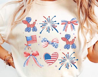 May include: Cream-colored t-shirt featuring a patriotic design. The design includes illustrations of fireworks, bows, a heart, and a balloon dog, all in red, white, and blue. An American flag is also featured.