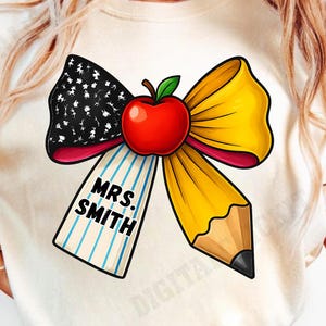May include: Cream-colored t-shirt featuring a bow design with an apple and pencil. The bow has a black and white pattern, yellow ribbon, and a red apple. The pencil is yellow and brown. The text "MRS. SMITH" is on a blue and white striped tag.