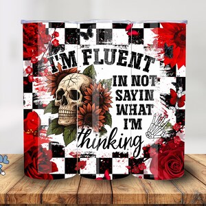 Könnte beinhalten: Ein Edelstahlbecher mit einem Totenkopf- und Sonnenblumen-Design. Der Becher trägt den Text "I'm fluent in not saying what I'm thinking" auf einem schwarz-weiß karierten Hintergrund mit roten floralen Akzenten und Schmetterlingen.