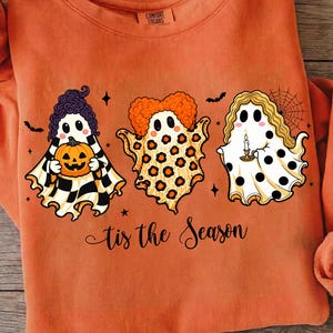 Puede incluir: Una sudadera naranja de manga larga con un gráfico de tres fantasmas con peinados y patrones distintos. Los fantasmas sostienen una calabaza, una vela y están junto a una telaraña. El texto 'tis the Season' está impreso en la parte inferior.
