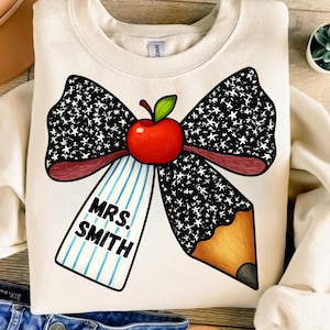 May include: Cream-colored sweatshirt featuring a large bow design with a red apple and pencil. The bow is black with white stars. A tag with the text "MRS. SMITH" hangs below the apple. The sweatshirt is a teacher-themed item.