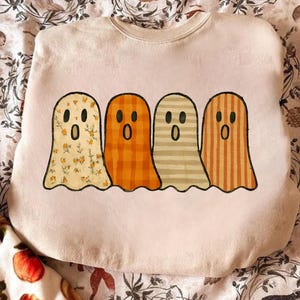 Op de afbeelding: Een lichtroze sweatshirt met vier cartoongeesten. Elke geest heeft een ander patroon: bloemen, ruitjes, strepen en verticale strepen. De geesten hebben zwarte ogen en open monden. De sweatshirt heeft een ronde hals.