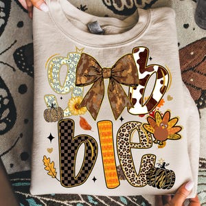 May include: Beige sweatshirt with the words "Grateful, Blessed, Thankful" in a fall-themed design. The design includes a large bow, pumpkins, leaves, and a turkey. The letters have various patterns and textures.