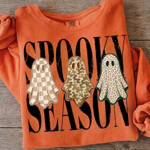 May include: An orange long-sleeve sweatshirt featuring the black text SPOOKY SEASON. Three ghost illustrations are printed across the front: one with a beige checkerboard pattern, one with a green camouflage print, and one with a leopard print.