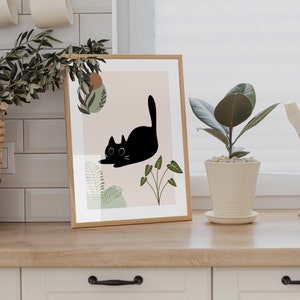 May include: A framed print with a black cat illustration and green foliage on a light pink background. The print is in a wooden frame and is sitting on a wooden countertop.