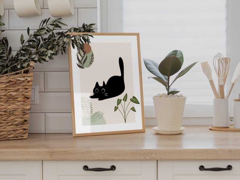 May include: A framed print with a black cat illustration and green foliage on a light pink background. The print is in a wooden frame and is sitting on a wooden countertop.