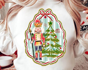 May include: White sweatshirt with a watercolour design of a nutcracker soldier and Christmas tree within a decorative frame. The design includes red, green, and gold accents, ideal for festive wear.