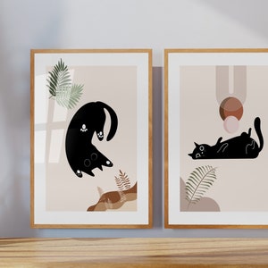 May include: Two framed prints with a minimalist design featuring black cats and abstract shapes in brown, pink, and beige tones. The prints have a light beige background and are framed in natural wood.